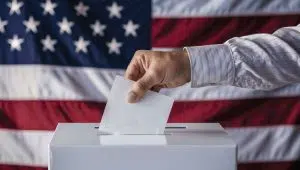 Hand putting a ballot in a ballot box with an American flag in the background.