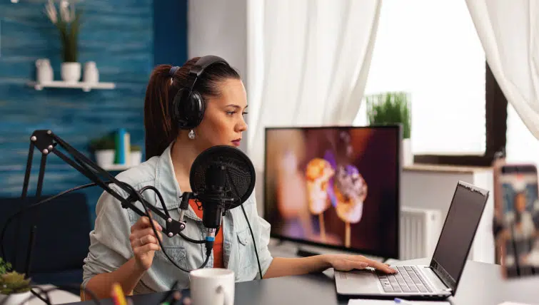 A young female social media influencer broadcasting herself with a laptop, microphone, and head set.